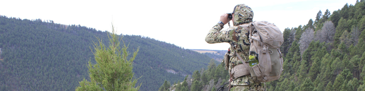 A person in full camouflage outdoor gear with a large backpack stands on a rocky hillside overlooking a forested mountain valley, holding binoculars to their eyes.